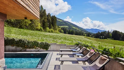 Liegestühle neben Pool mit Berg- und Wiesenblick bei Sonnenschein