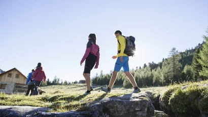 Vier Personen wandern auf einem sonnigen Bergweg mit Häusern und Wäldern