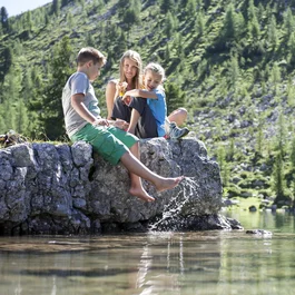 Drei Kinder sitzen am Ufer eines klaren Bergsees und plantschen im Wasser